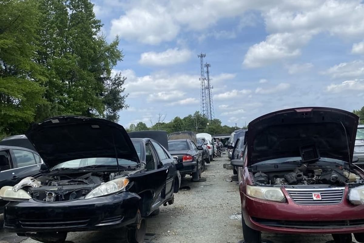 Pick Your Part - Greensboro: Rows of salvage cars with open hoods on gravel in a self-service junkyard under a partly cloudy sky