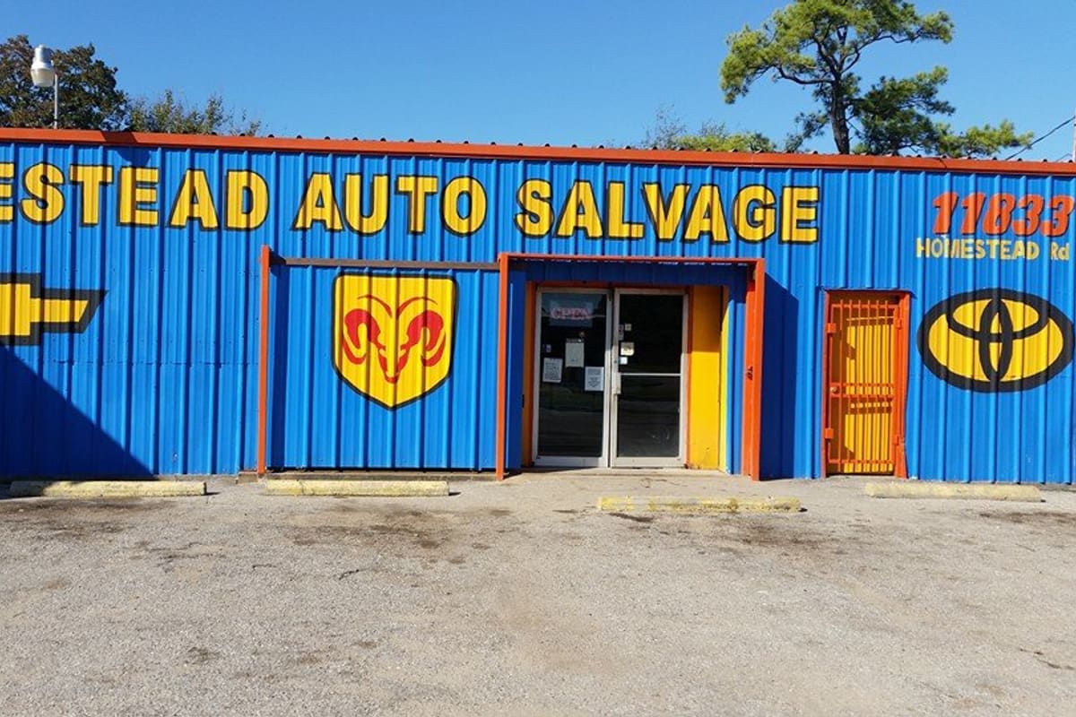 Homestead Car and Truck Parts: Blue metal storefront with yellow lettering, car brand logos, a glass entrance door and an open sign above a gravel lot