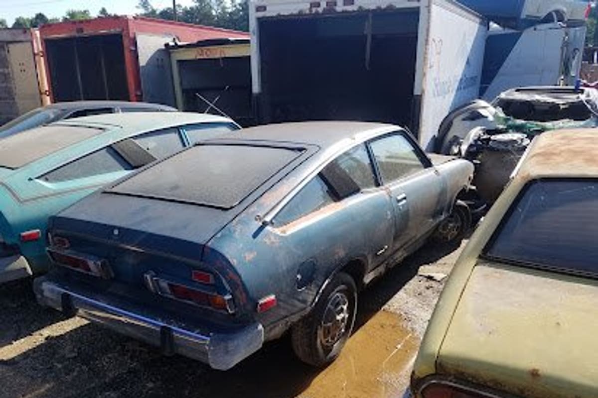 Wheeley Auto Mall: Rusty vintage cars parked in a salvage yard beside box truck trailers on a sunny day