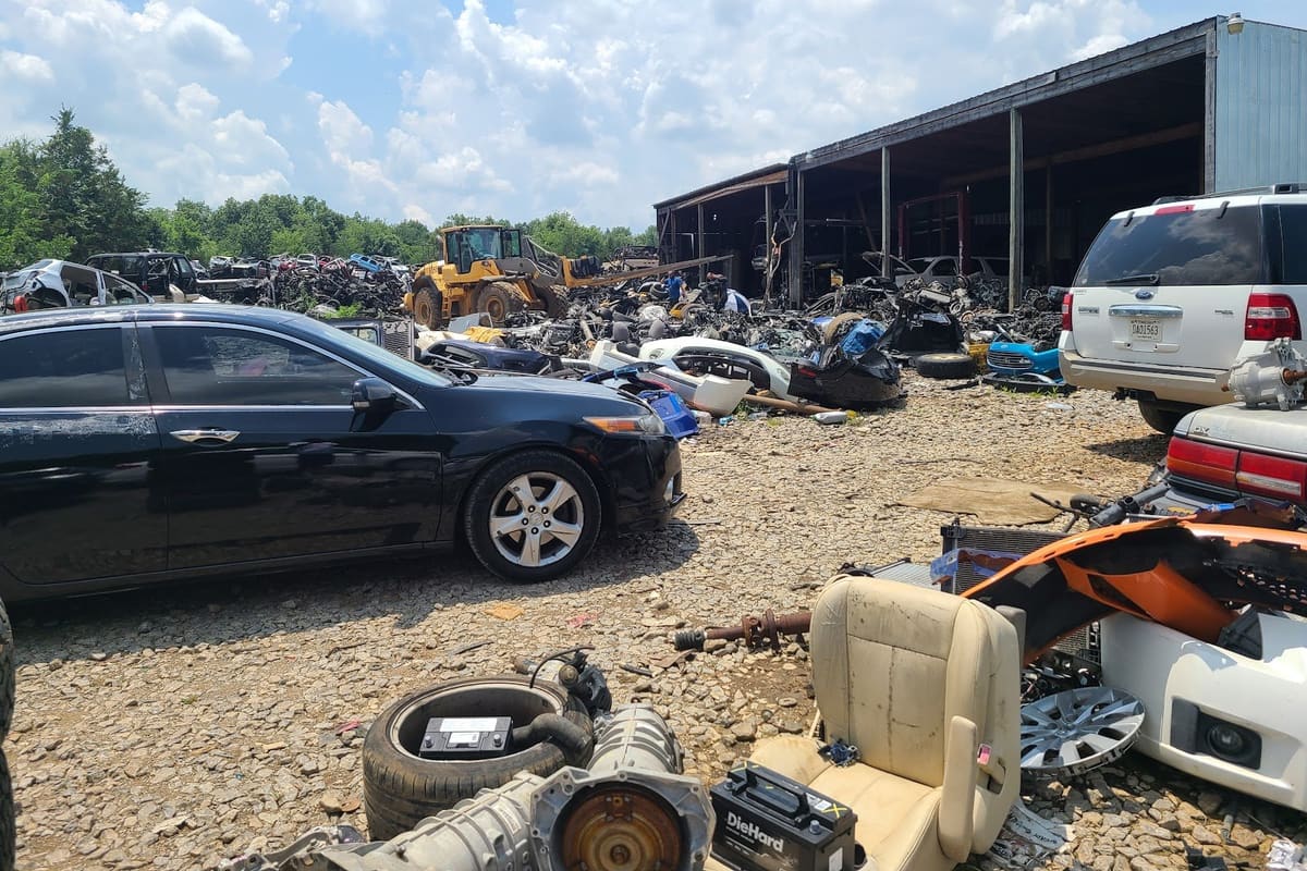 Manus Road Auto Salvage: Wide daytime view of a gravel salvage yard with cars, loose parts, a front loader and an open-sided storage shed