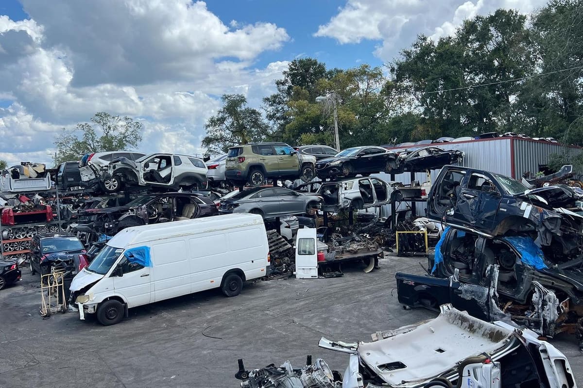 Oscar Used Auto Parts: Wide view of a salvage yard with stacked wrecked cars, a white van, and vehicle parts on a paved lot under a cloudy sky