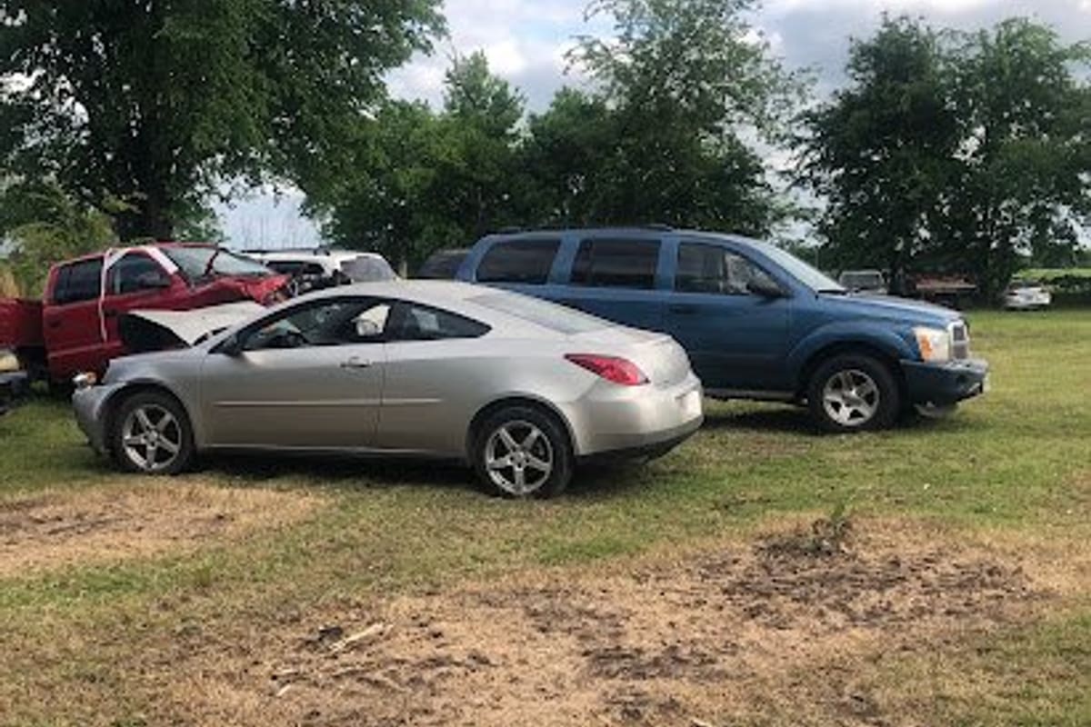 Mora's Recycling: Several salvage vehicles parked on a grassy lot with trees in the background, including a silver coupe and a blue SUV