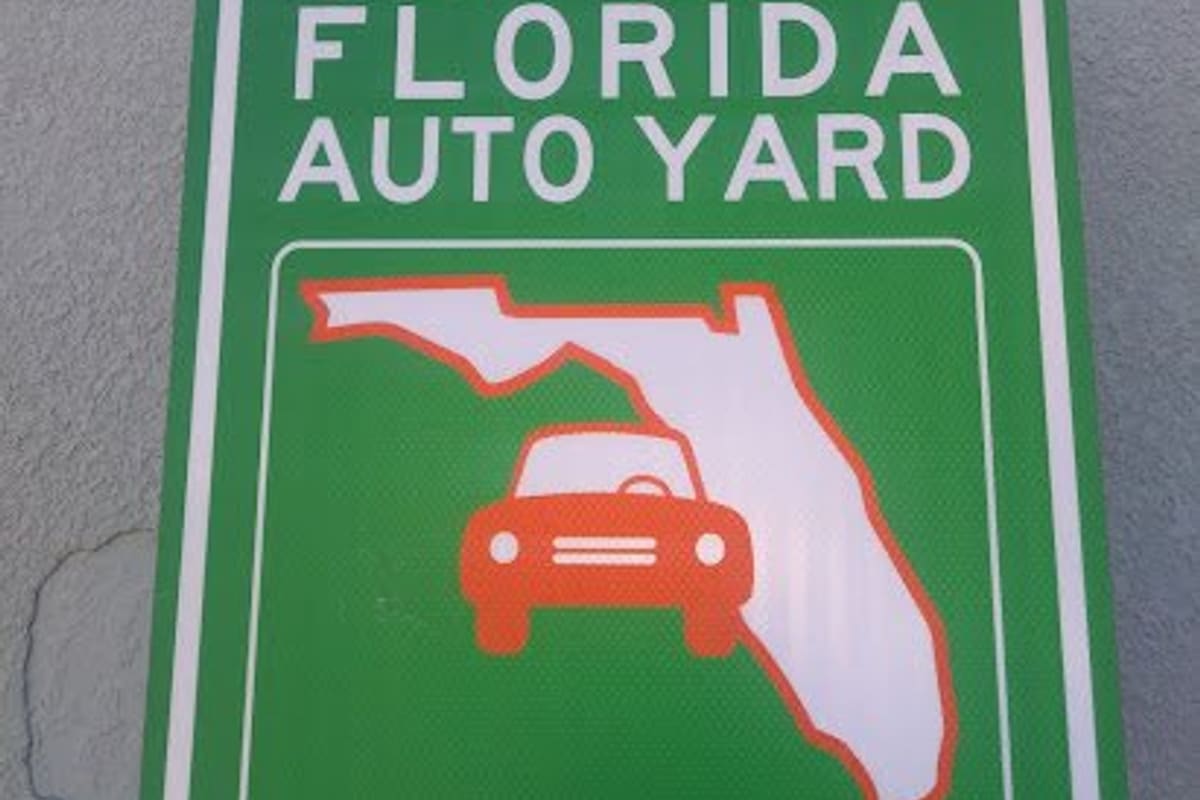 Central Florida Auto Yard: Green road-style sign with a car on a Florida map, listing the address, phone number and hours, mounted on a stucco wall