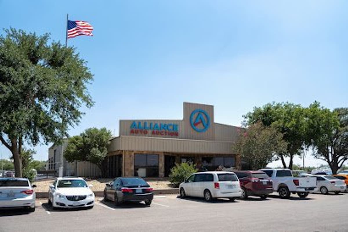 Alliance Auto Auction Abilene: Tan commercial building with a large blue sign and flag, with cars parked in the front lot on a clear day