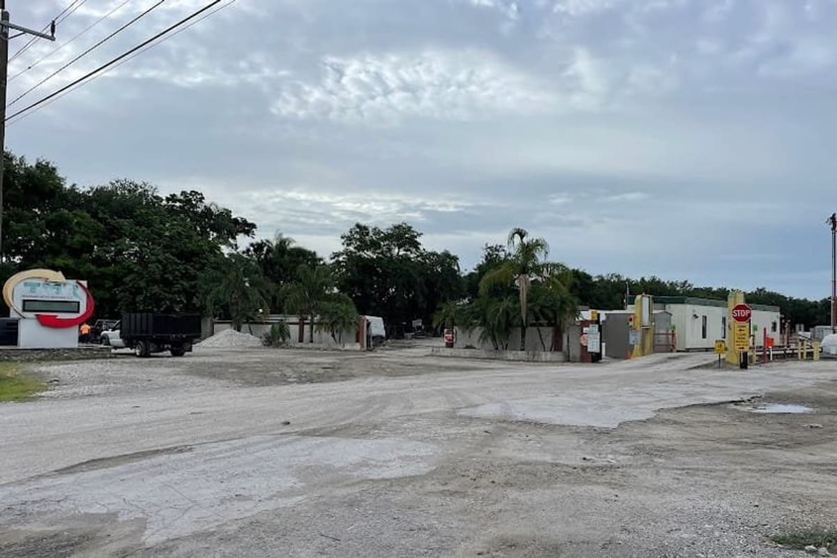 Trademark Metals Recycling: Gravel entrance road leading to a scrap yard with a stop sign, small office buildings, palm trees and a sign at left