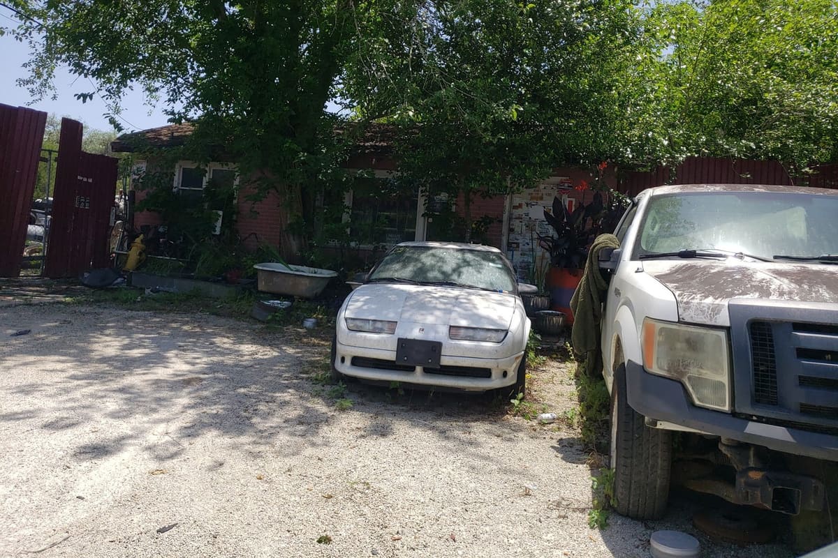 John's Pull & Save: Gravel lot with a white car and a pickup truck parked in front of a brick building shaded by large trees