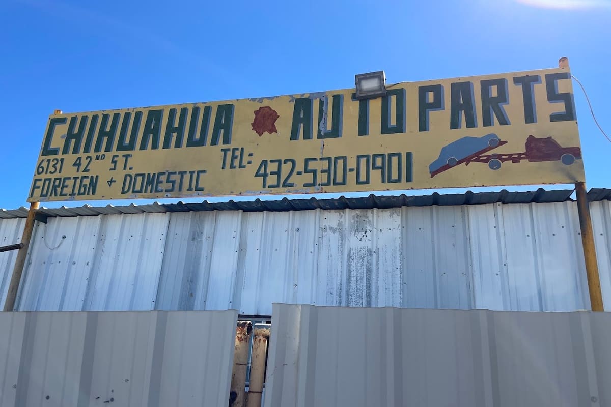 Chihuahua Auto Parts: Hand-painted yellow auto parts sign with phone number above a corrugated metal fence under a clear blue sky