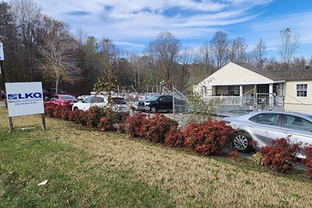 LKQ Salisbury: Auto salvage yard with parked cars behind red shrubs, roadside signs, and a small office building