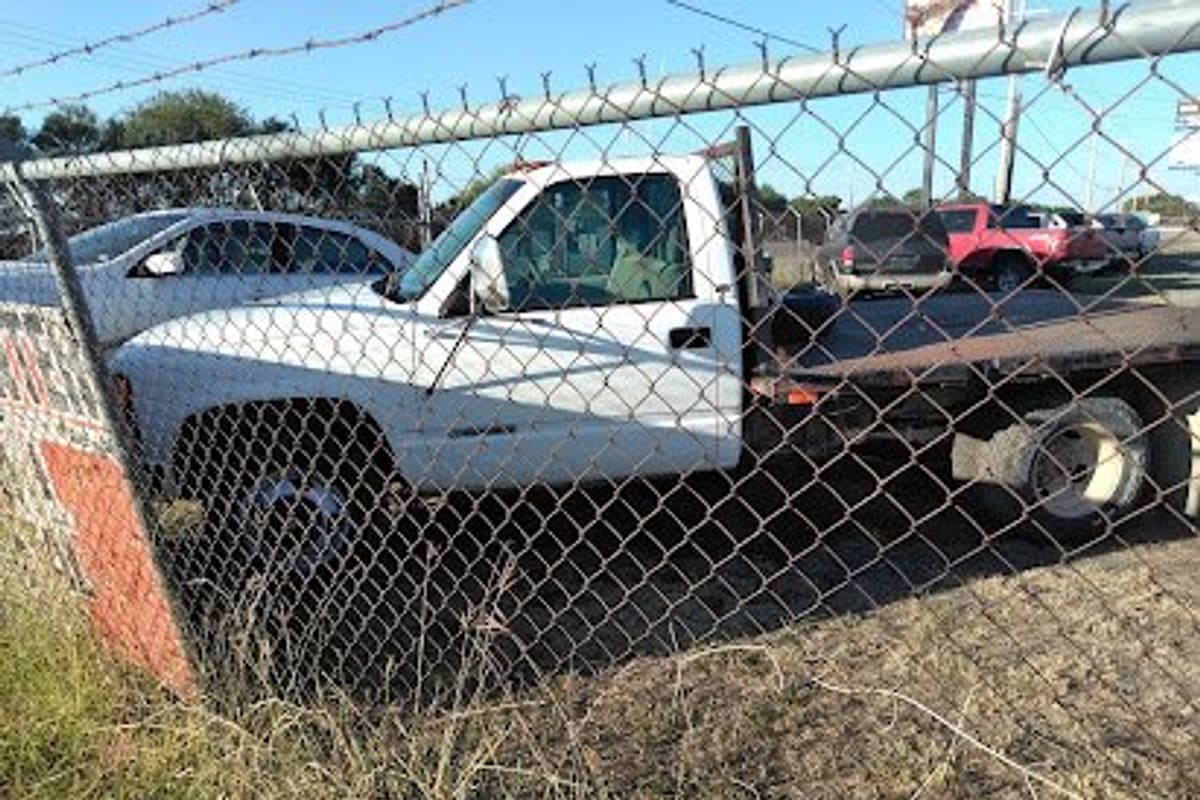 Hacienda Salvage and Metal: White flatbed tow truck and other vehicles parked in a dirt lot seen through a chain-link fence on a sunny day