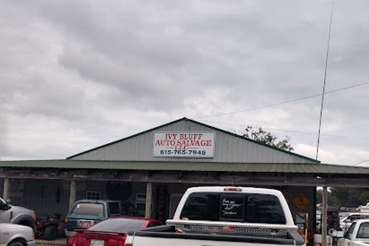 Ivy Bluff Auto Salvage: Covered front of a gray metal salvage office building with a sign, trucks and cars parked on gravel in front