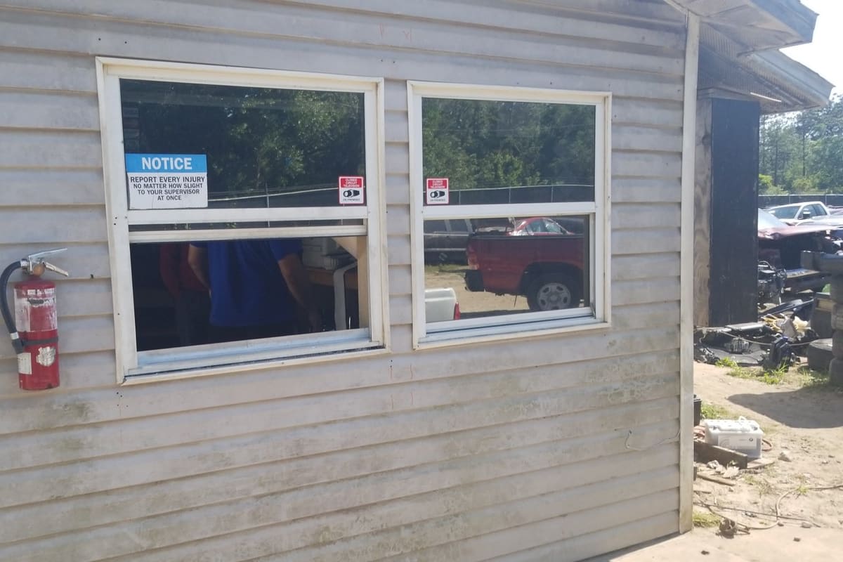 Energy Auto Dismantling: Weathered gray siding office building with a vertical OFFICE sign, two windows, and a fire extinguisher beside a salvage yard