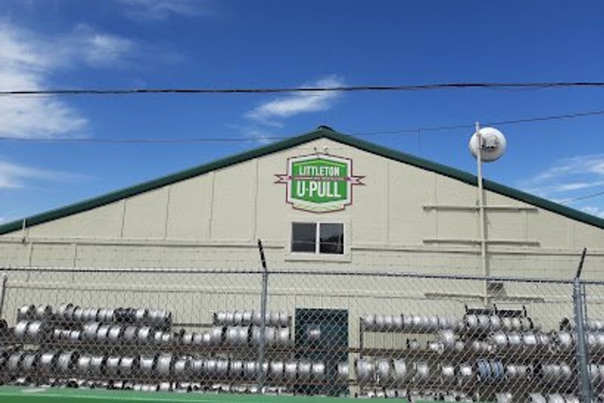 Littleton U-Pull: Tan metal building with a green gabled roof and a green U-Pull sign, with stacked wheel rims behind a chain-link fence