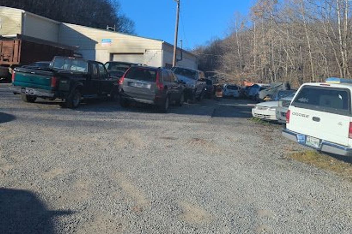 Goodlettsville Auto Salvage: Gravel lot with parked trucks and cars in front of a low warehouse building, with wooded hills behind under a clear blue sky