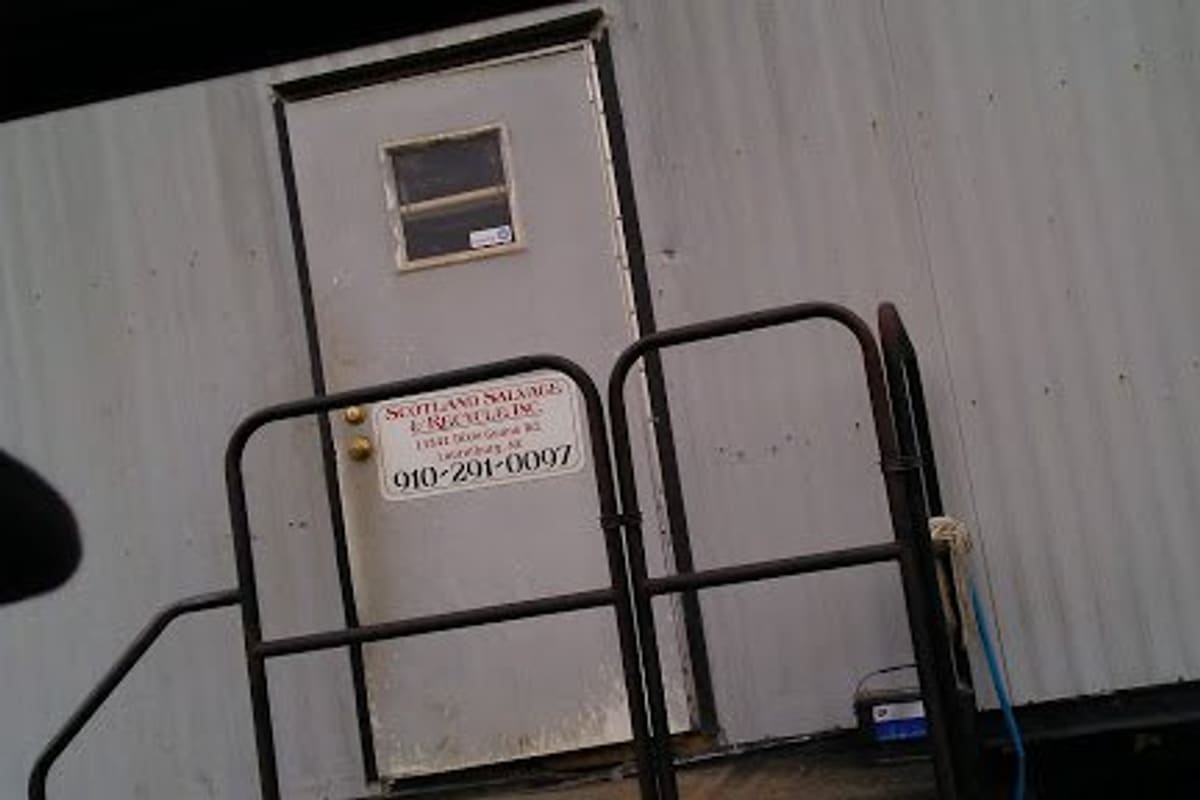 Auto Recyclers of Laurinburg: Metal-sided office trailer with a white door, a small posted sign with a phone number, and black railings in front