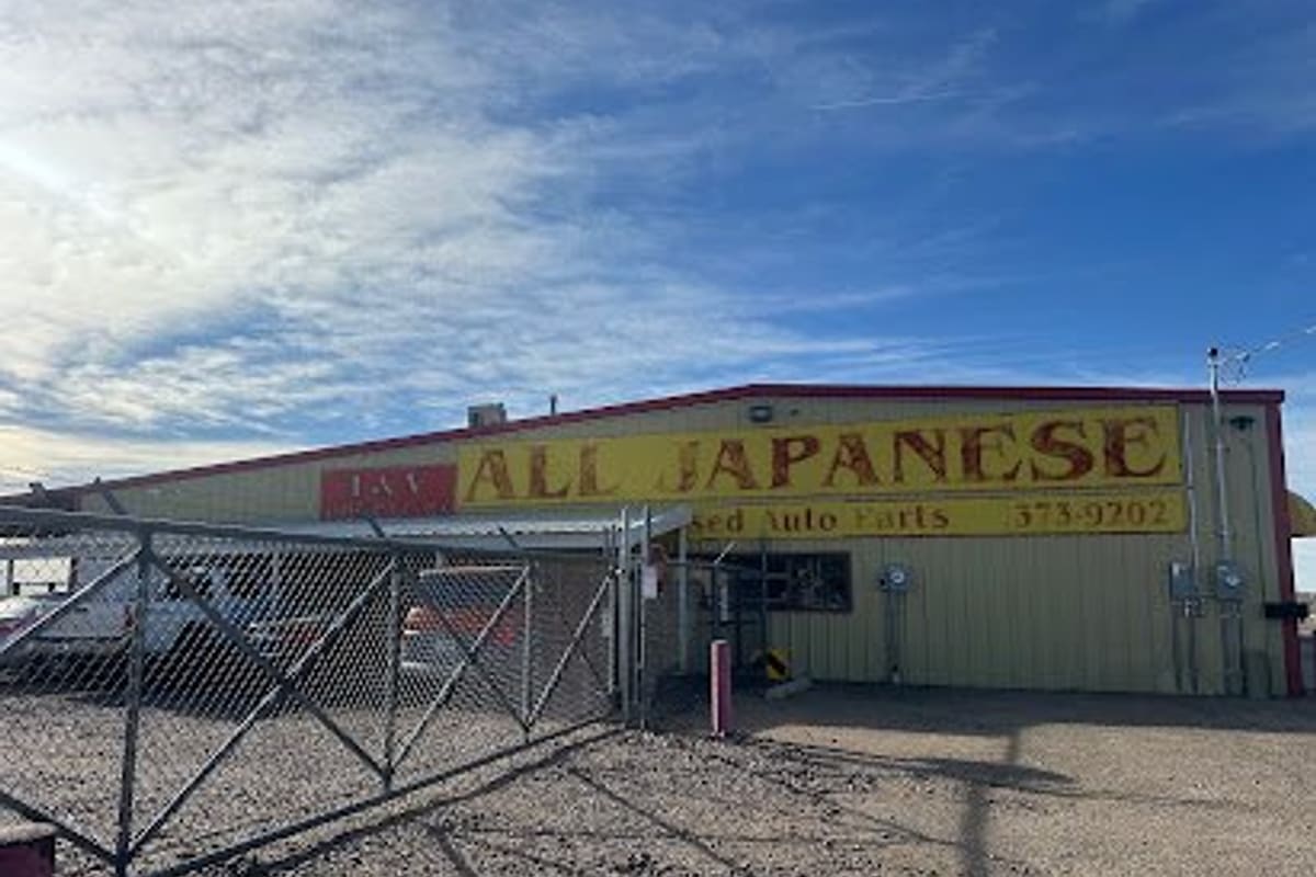 All Japanese Used Auto Parts: Yellow metal warehouse building with a large painted sign for used Japanese auto parts, behind a chain-link gate and gravel lot