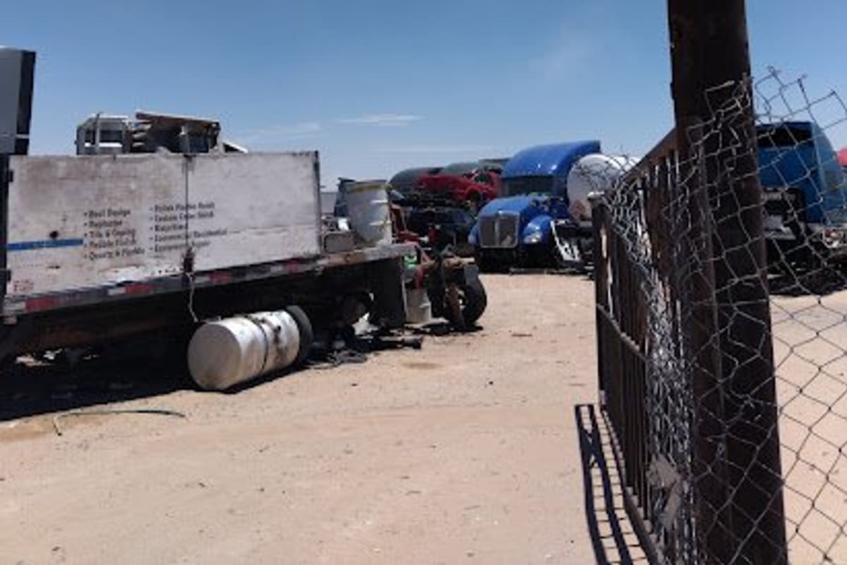 Miky's Truck Parts Sales: Dirt salvage yard with a flatbed truck, several semi truck cabs and a chain-link gate under a clear blue sky