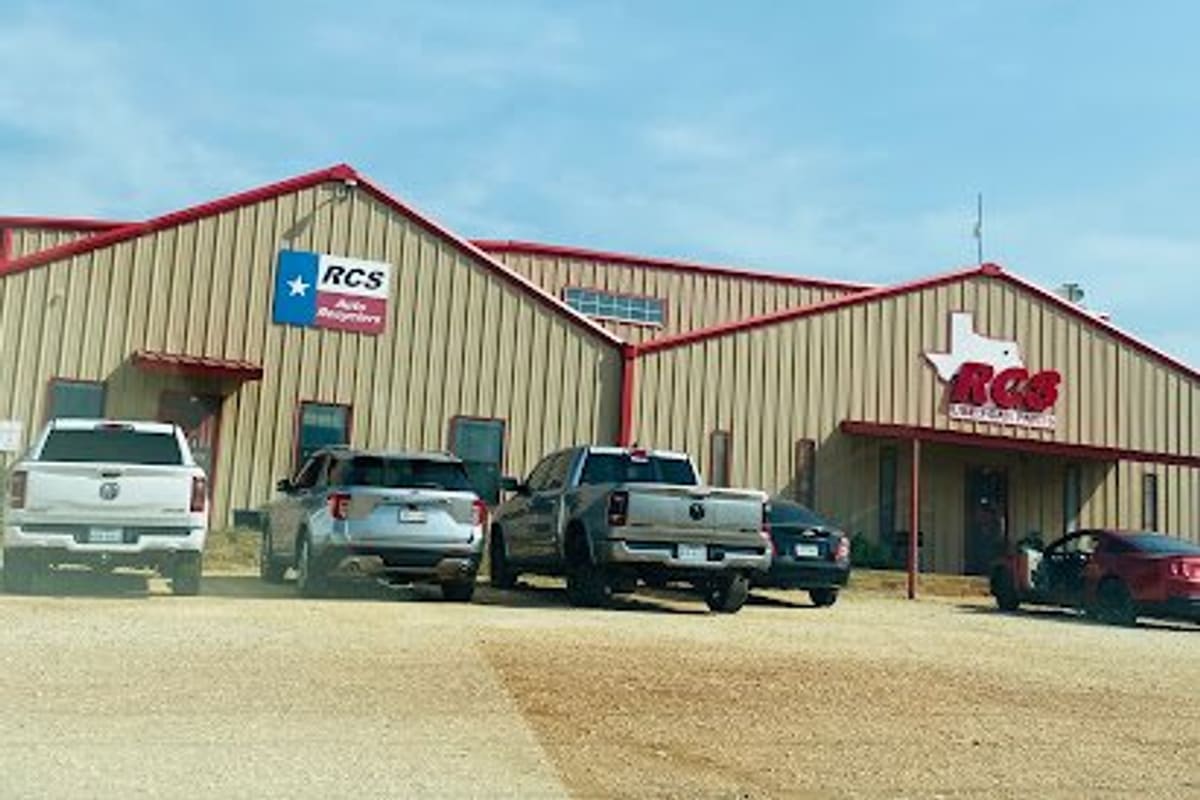 RCS Auto Recyclers: Tan metal building with red trim and Texas-themed signs, with pickups and cars parked on a gravel lot under a blue sky