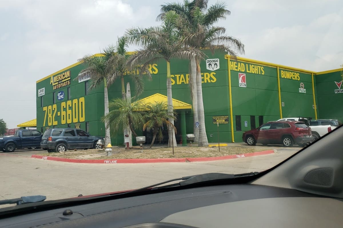American Used Auto Parts: Green and yellow auto parts building with palm trees, a phone number on the wall, and cars parked out front