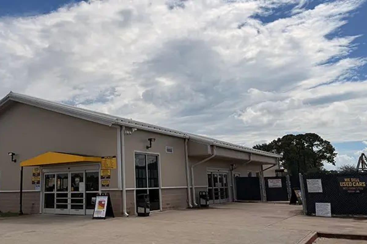 U-Pull-&-Pay Houston: Tan single-story office building with yellow entrance awning beside a fenced yard gate under a cloudy sky