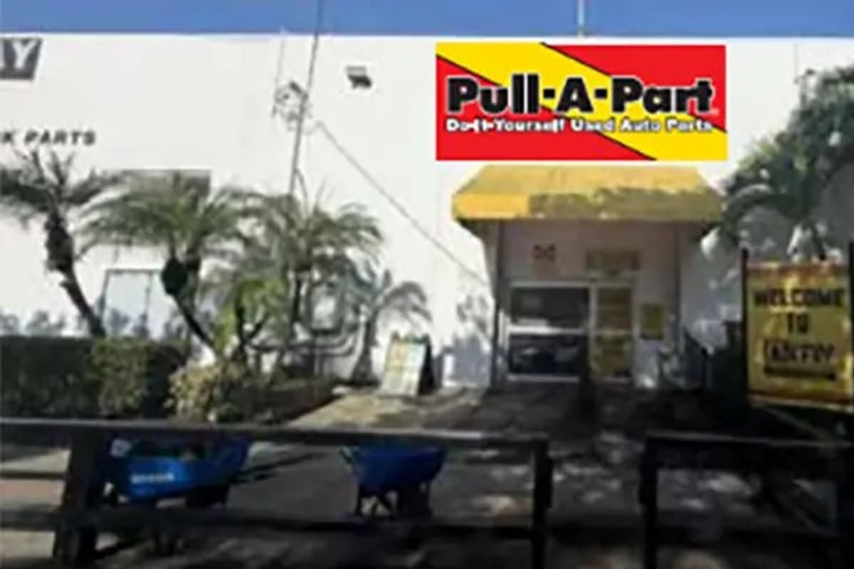 Pull-A-Part West Palm Beach: Entrance to a white auto parts building with a yellow awning, palm trees, and a welcome sign by the gate