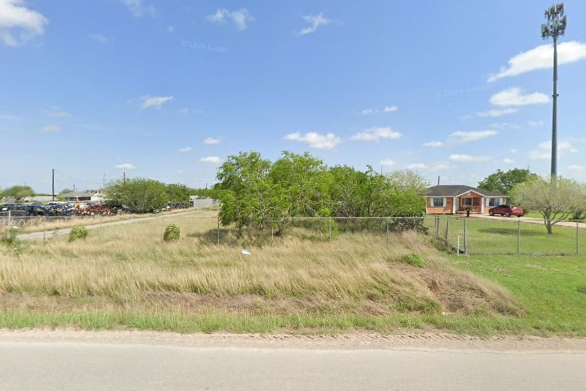 Estrada Recycling: Street view of a fenced grassy lot with parked vehicles in the distance and a house nearby under a blue sky