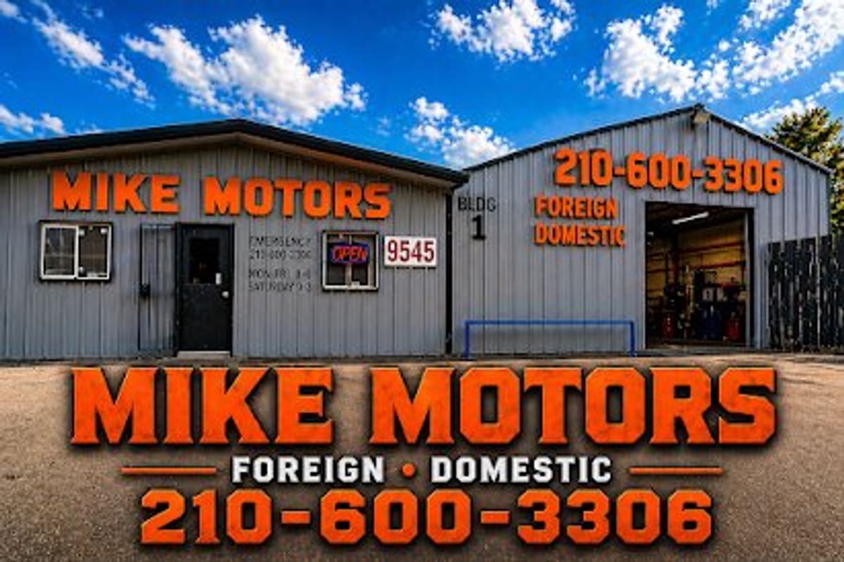 Mike Motors Auto Salvage: Gray metal shop buildings with orange lettering, an open bay door and a black entrance door under a blue sky