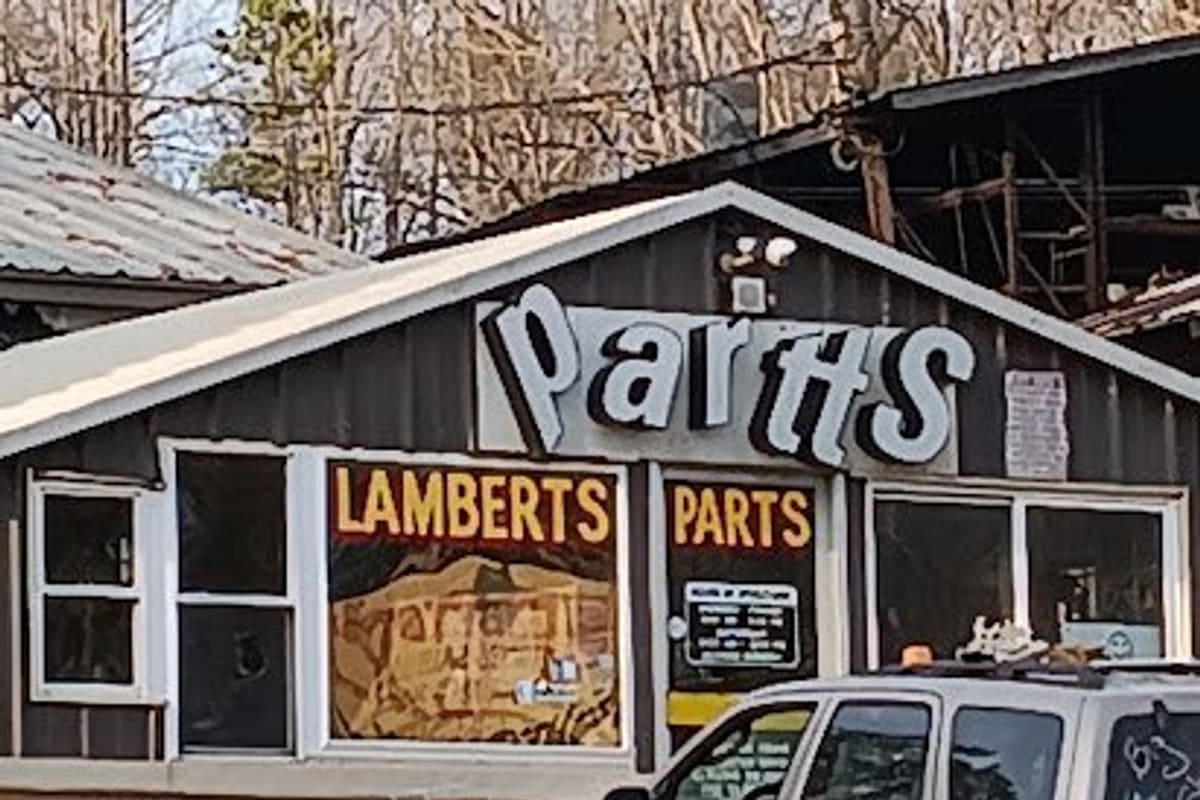 Lambert Auto Parts: Small dark-sided shop building with a large white parts sign, painted window signs and a silver SUV parked in front