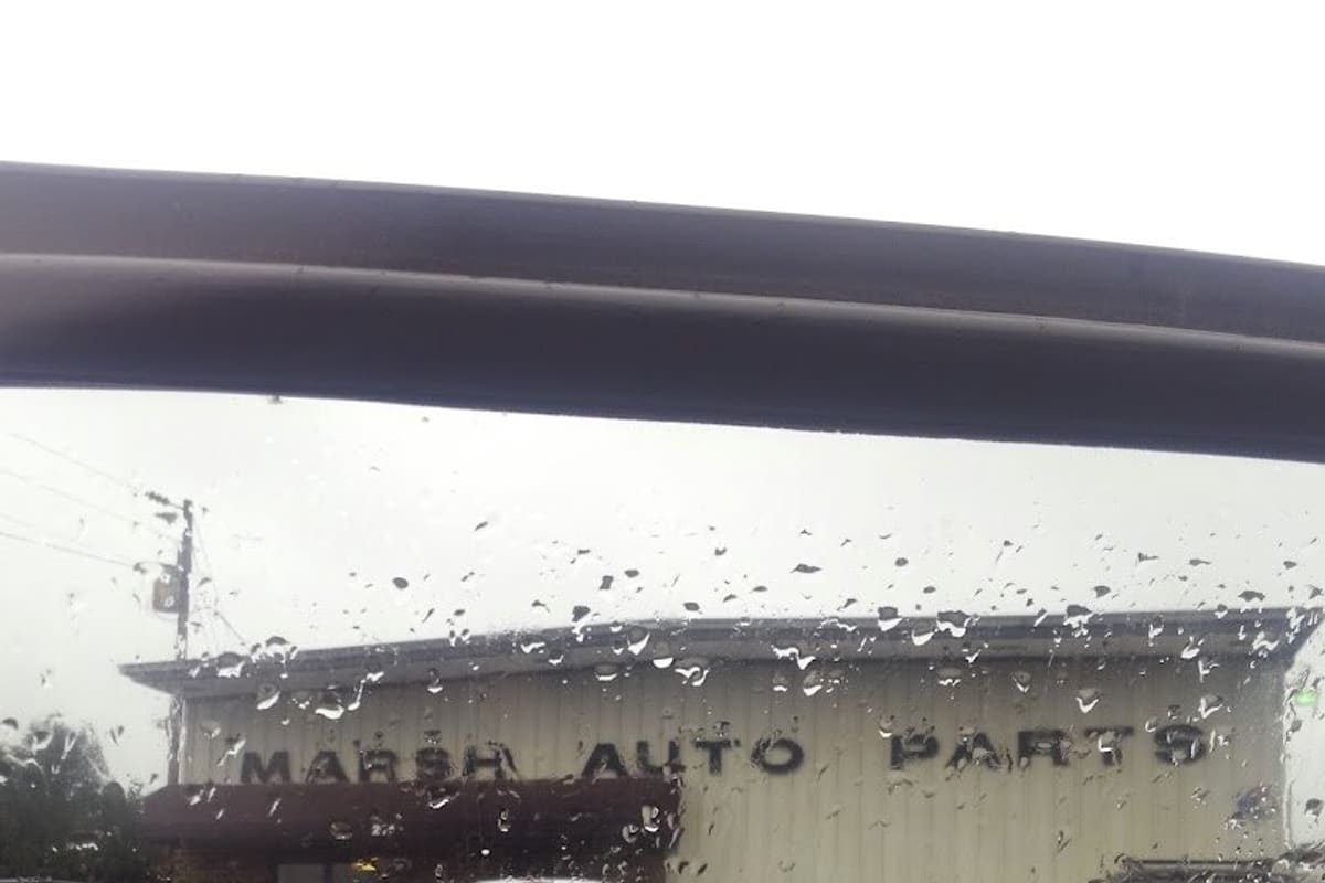 Marsh Auto Parts-Siler City: Rain-spattered view of a tan metal auto parts building with lettering on the wall and trucks parked in the wet lot