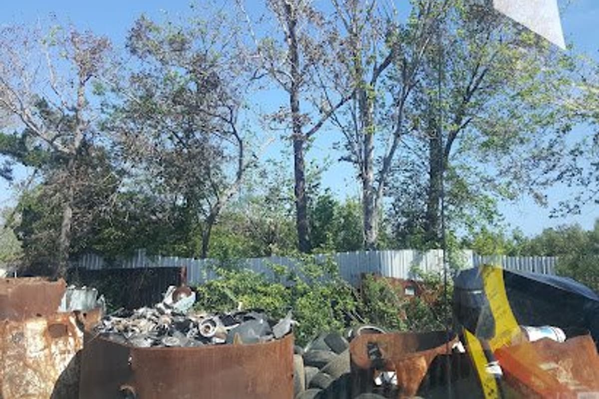 Holly Hill Salvage & Junk: Rusty barrels filled with scrap metal and tires in a salvage yard, with a metal fence and trees behind