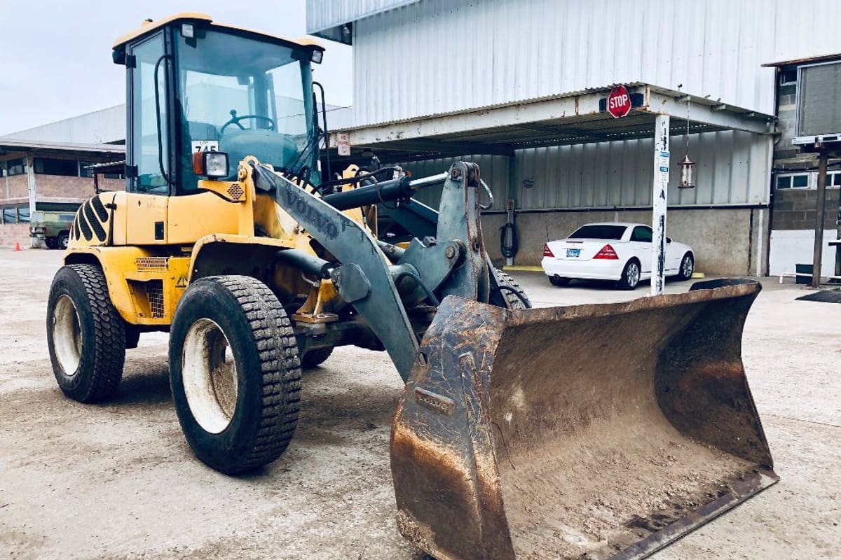 San Antonio Truck & Equipment: Yellow wheel loader with a dirty bucket parked on a gravel lot in front of metal-roofed buildings and a parked white car