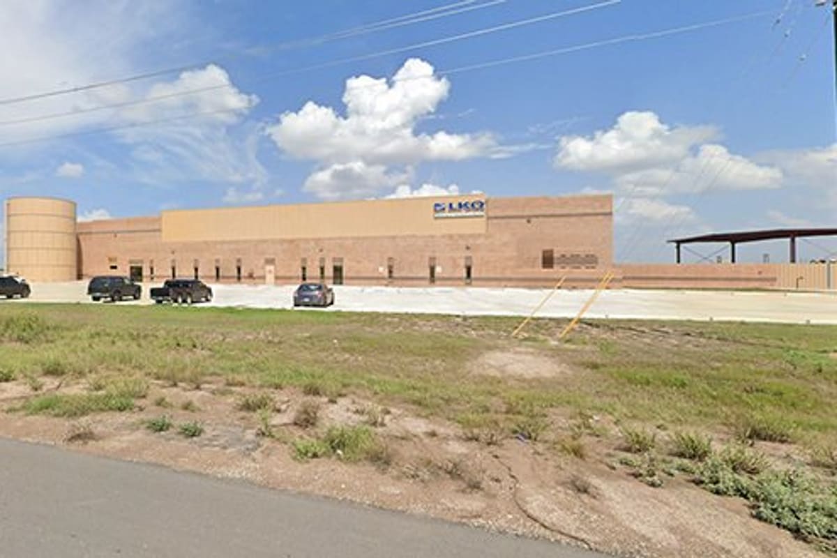 LKQ McAllen: Large tan brick industrial building with parked trucks in front, seen across a grassy field from the road