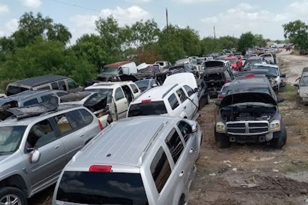 Gomez Junk Yard: Rows of salvage cars and SUVs parked on dirt ground under a partly cloudy sky, with trees in the background