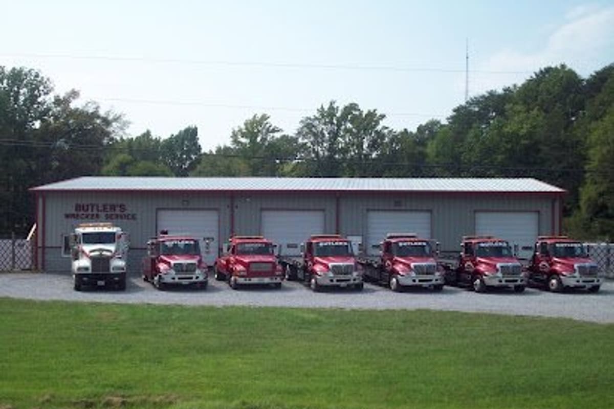 Butler Wrecker Service: Row of red and white tow trucks parked in front of a gray metal garage building with several bay doors, beside a grassy lawn