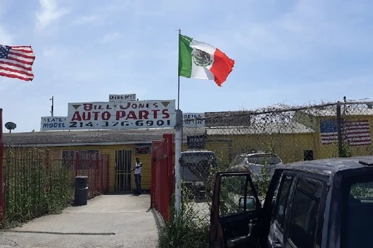 Bill Jones Auto Parts & Perez Used Cars: Yellow shop building with a hand-painted auto parts sign behind a fenced entrance, with American and Mexican flags flying