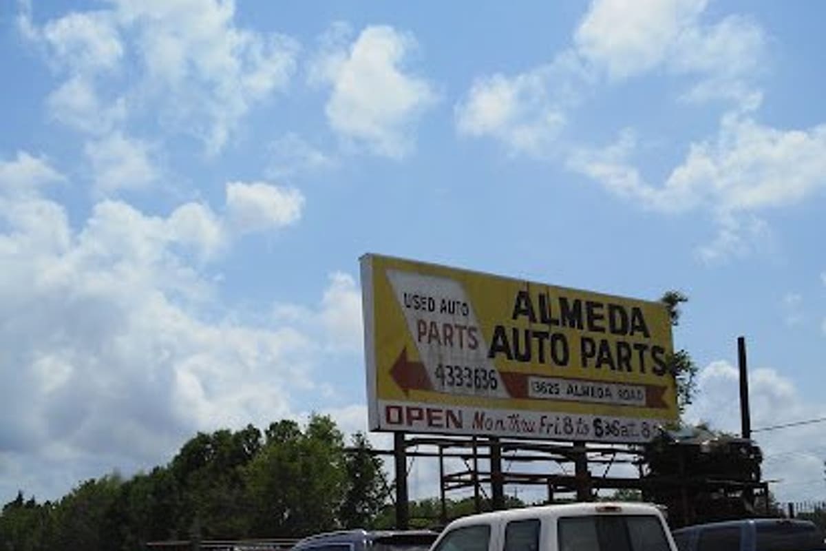 Almeda Auto Parts: Gravel lot with a white pickup and parked cars beneath a large yellow billboard sign for used auto parts
