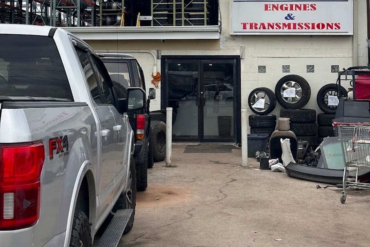 B &T Auto Parts: Tan building with a red and blue Engines & Transmissions sign, glass double doors, stacked tires and a parked pickup truck in front