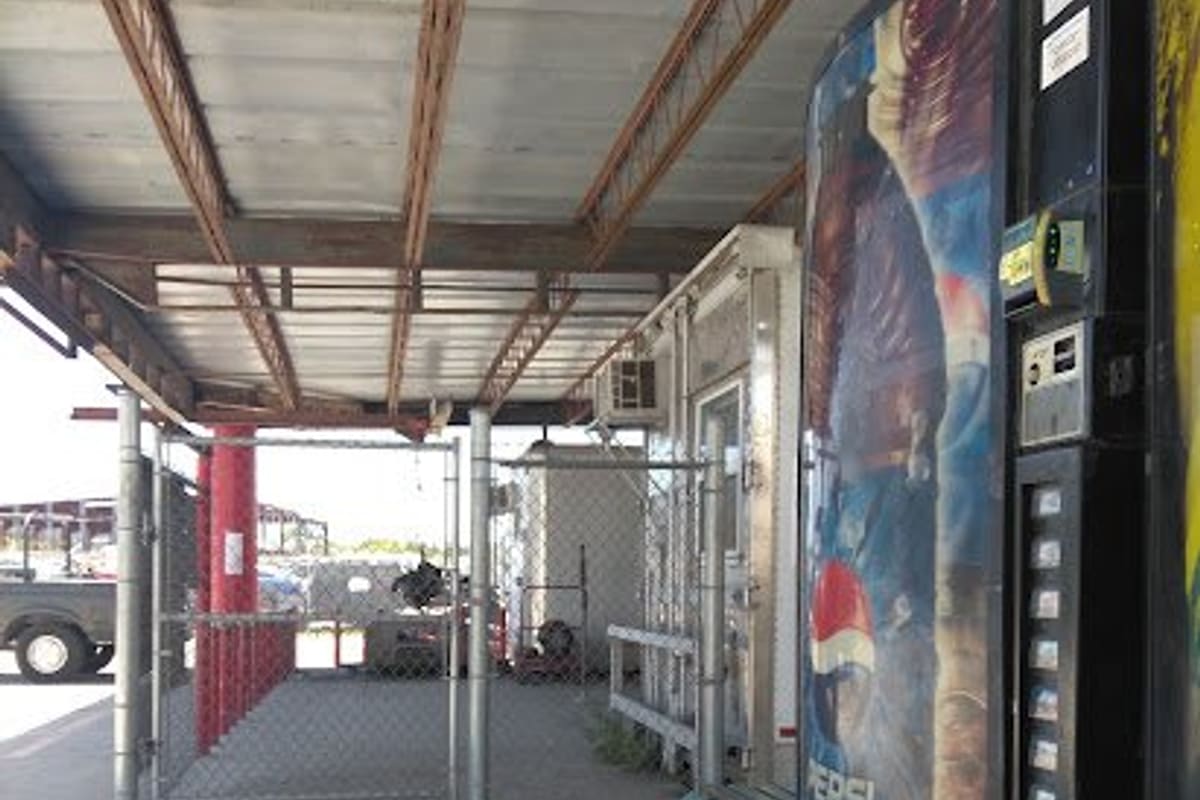 Gluecks Auto Parts: Covered walkway with a Pepsi vending machine, bench and chain-link gate beside a small office trailer