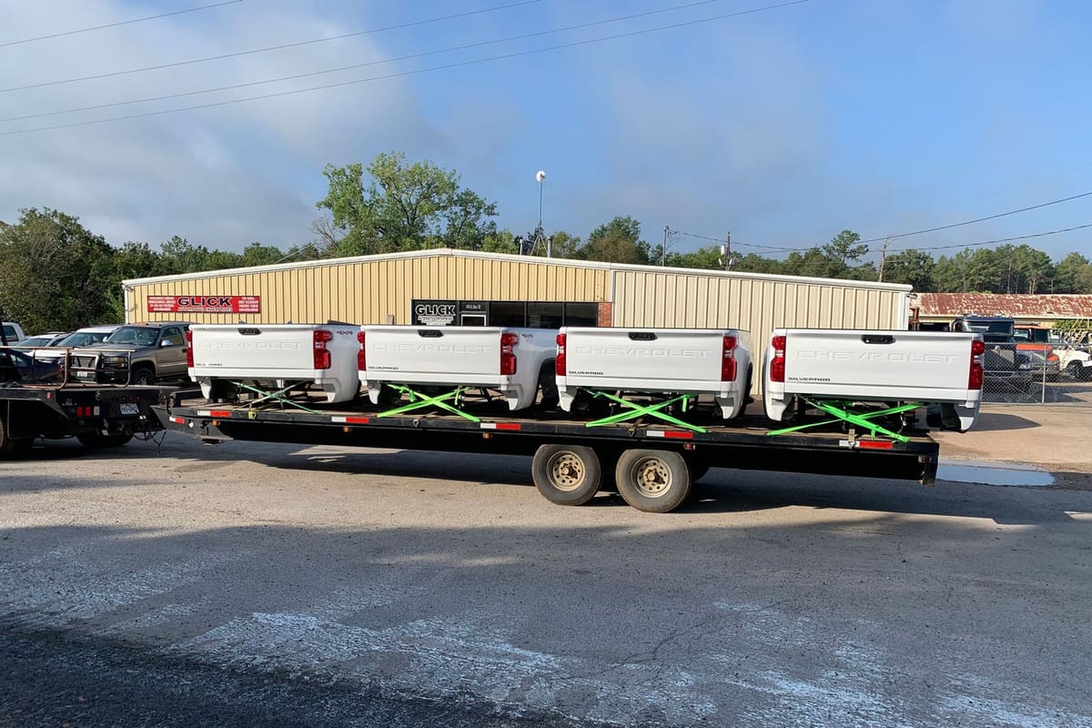 Glick Automotive Ent Nto Buffalo TX: Flatbed trailer loaded with white pickup truck beds parked in front of a tan metal shop building with a sign