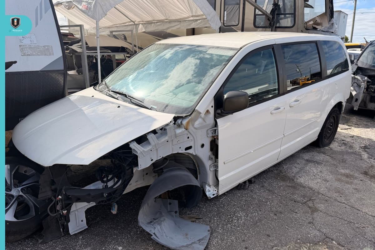 Doctor Parts: White minivan with its front end stripped sitting in a salvage yard beside a tent, trailers and other vehicles