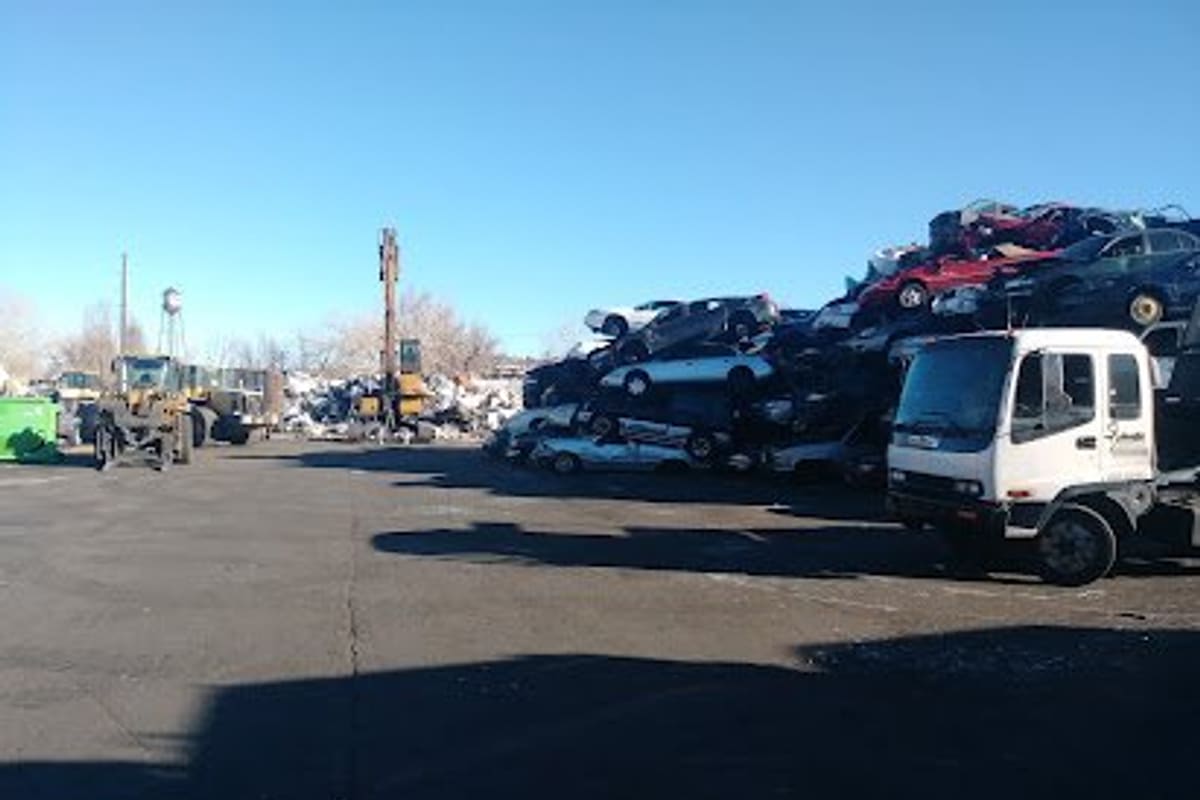 Denver Scrap Metal Recycling: Wide daytime view of a salvage yard with stacked crushed cars, a flatbed truck, and loaders on a paved lot under a clear sky