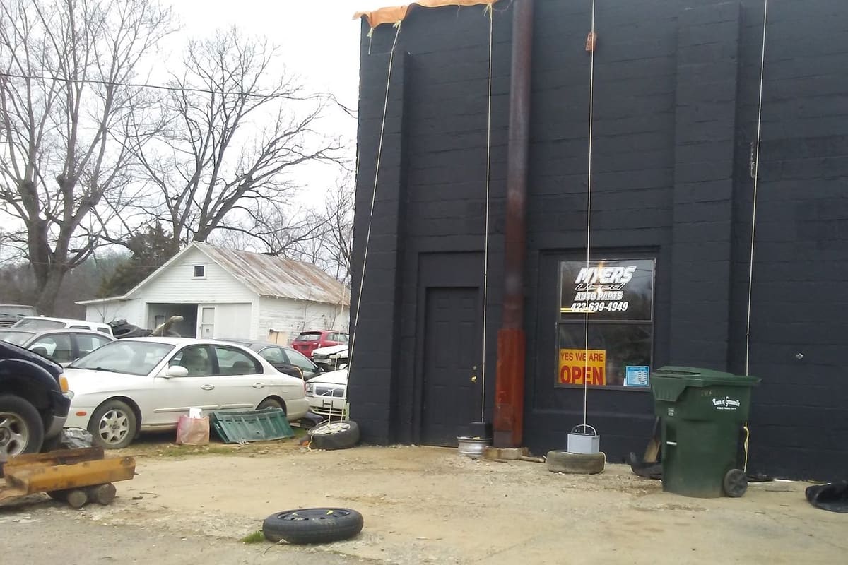 Myers Used Parts: Black concrete block shop building with a lit open sign in the window, cars parked alongside and a trash bin out front