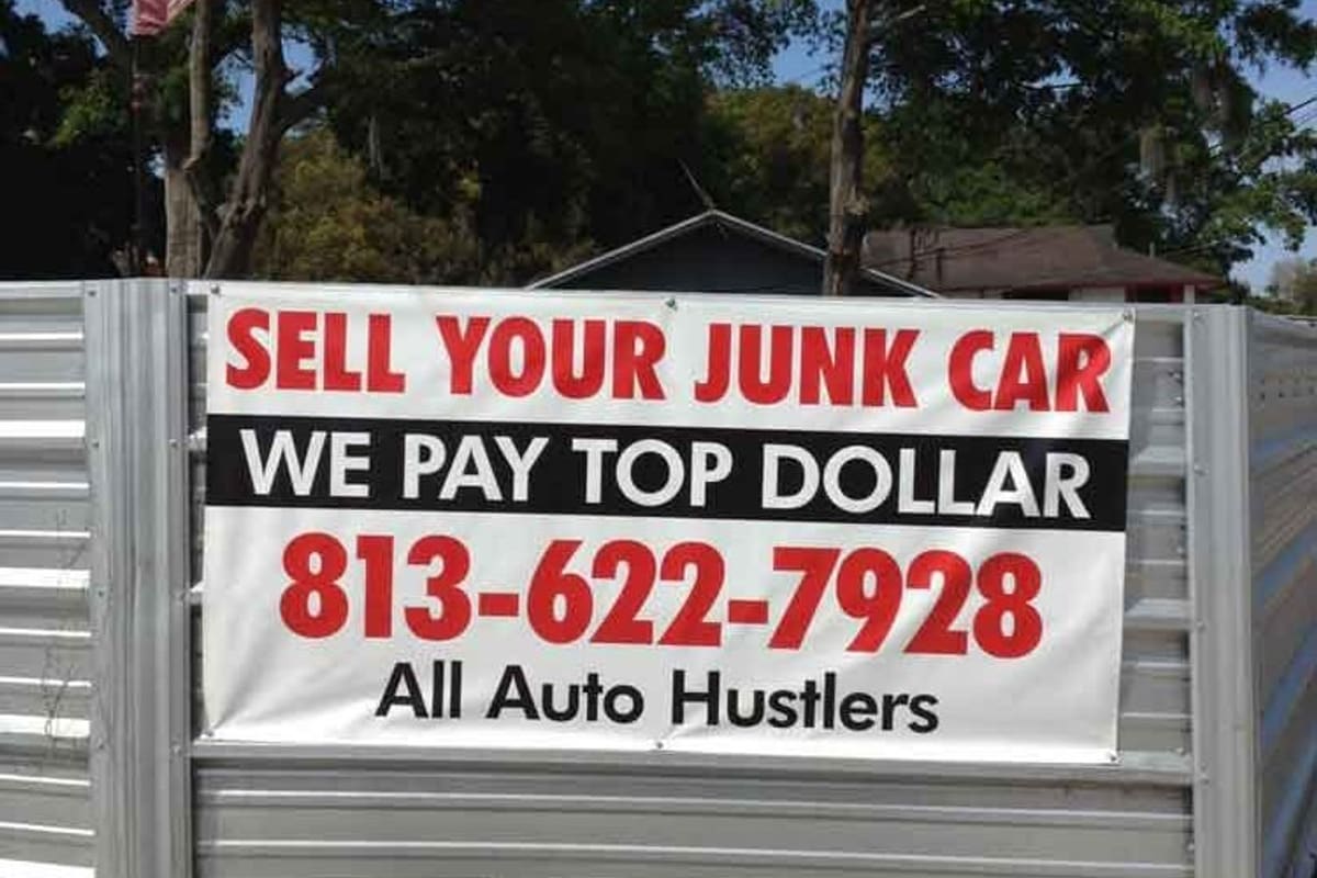 All Auto Hustler's: Banner on a corrugated metal fence reading Sell Your Junk Car, We Pay Top Dollar, with a phone number