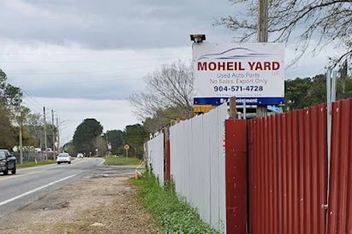 Moheil Yard: Roadside sign for a used auto parts yard on a fence of red and gray corrugated metal beside a two-lane road
