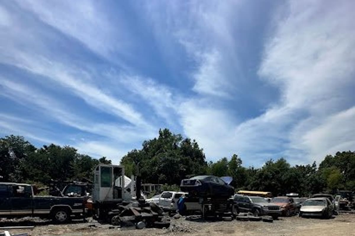Lebanon Scrap Metals & Parts: Dirt salvage yard with tire tracks, stacked cars, a pickup truck and heavy equipment under a blue sky with trees behind