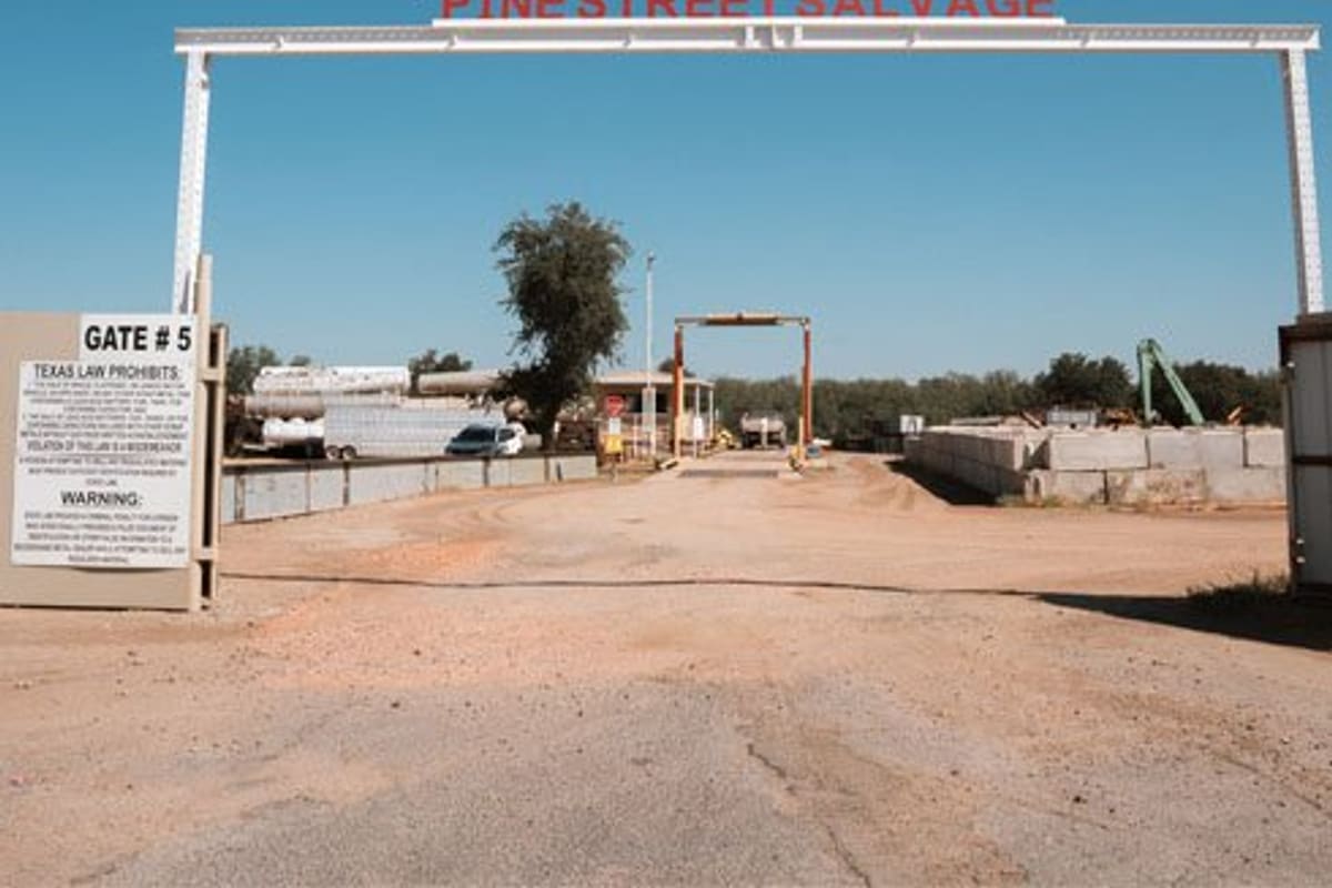 Pine Street Salvage: Overhead gate sign over a dirt entrance road leading into a salvage yard under a clear blue sky