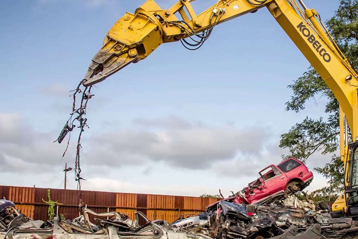 Patron Metal Recycling: Yellow excavator with shear attachment lifting wiring above a pile of crushed cars in a scrap yard