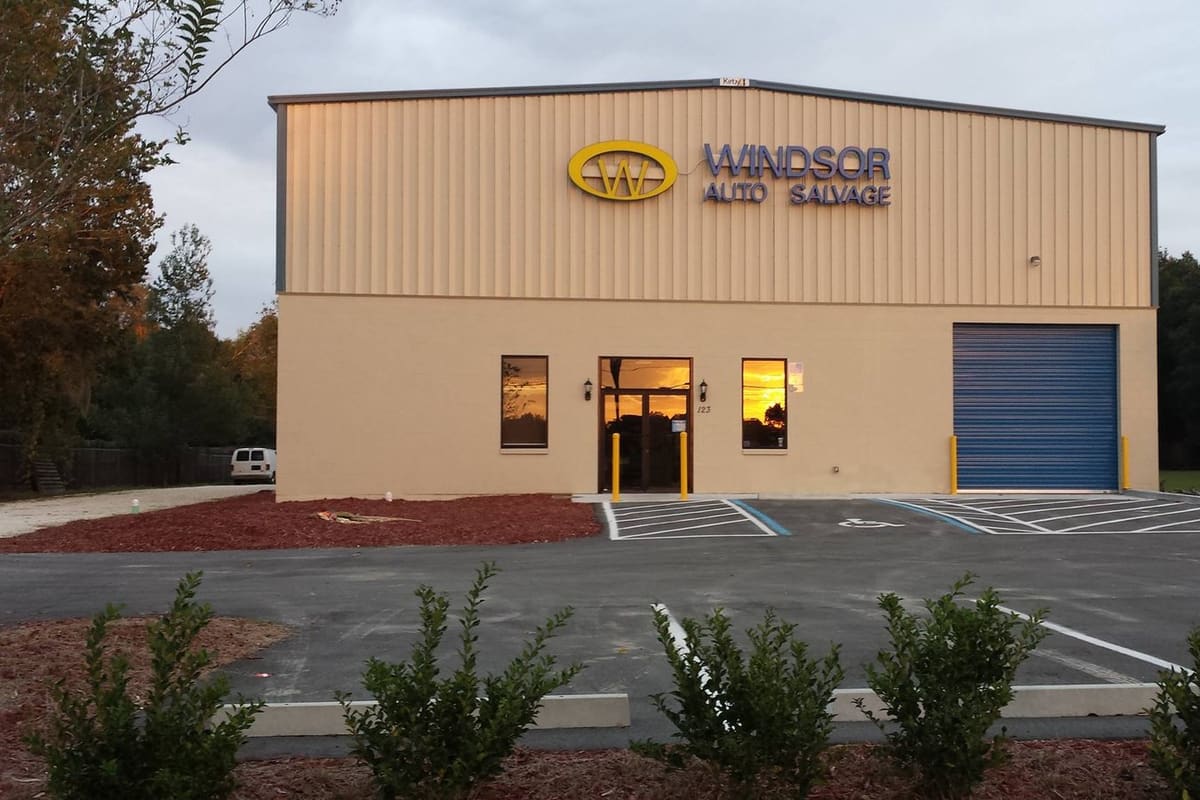Windsor Auto Salvage: Tan metal building with a gold and blue sign, glass entrance, blue roll-up door and paved parking lot at dusk