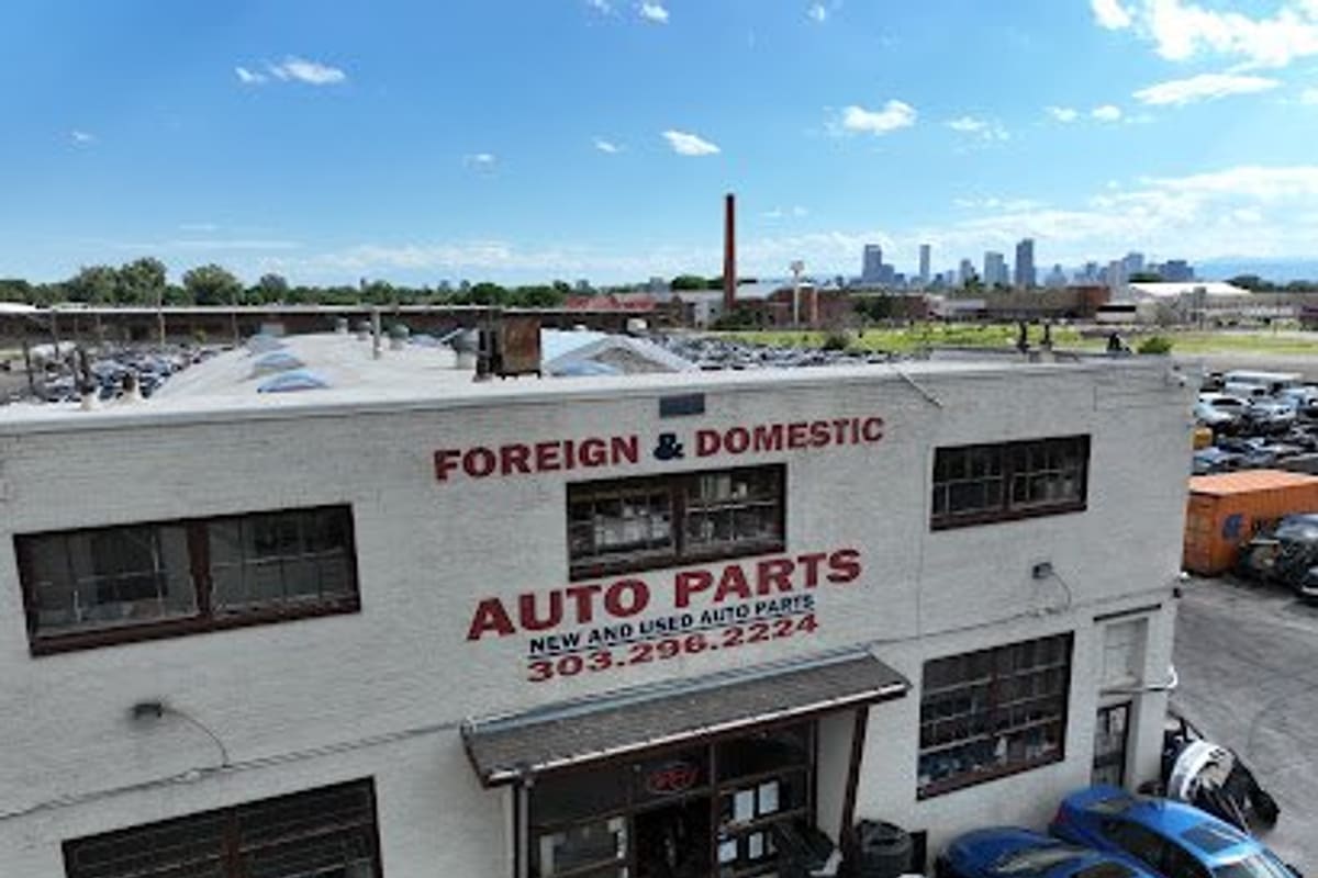 Foreign Used Auto Parts: Two-story white brick auto parts building with painted sign, awning entrance, tires and cars out front, city skyline behind