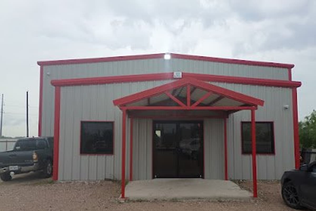 Mac Auto Parts: Gray metal building with red trim and a covered entrance, gravel lot in front with a pickup truck and car parked nearby