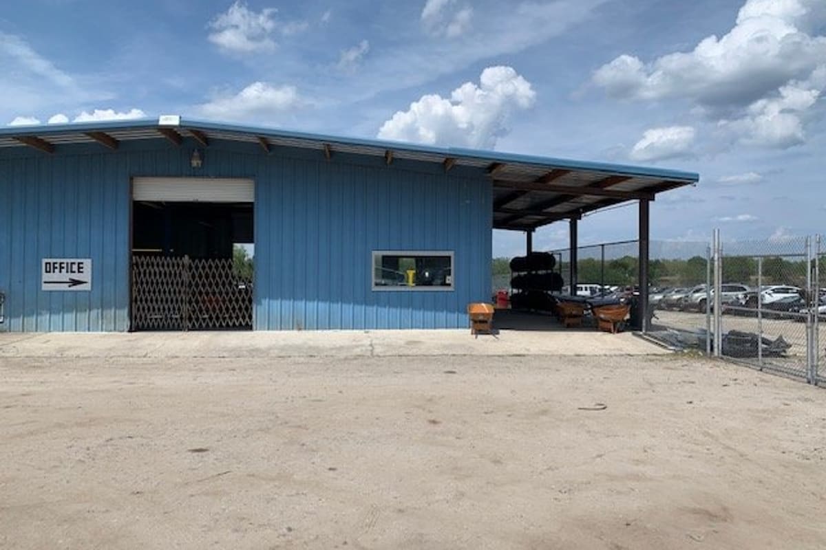 Marion County Auto Parts and Salvage: Blue metal shop building with a covered side bay, an office sign with arrow, and a fenced lot of cars beyond a dirt yard